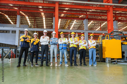 Diverse team of factory workers, engineers, and office staff pose confidently inside modern steel roofing sheet production plant. Their stance reflects unity, pride, and industrial teamwork