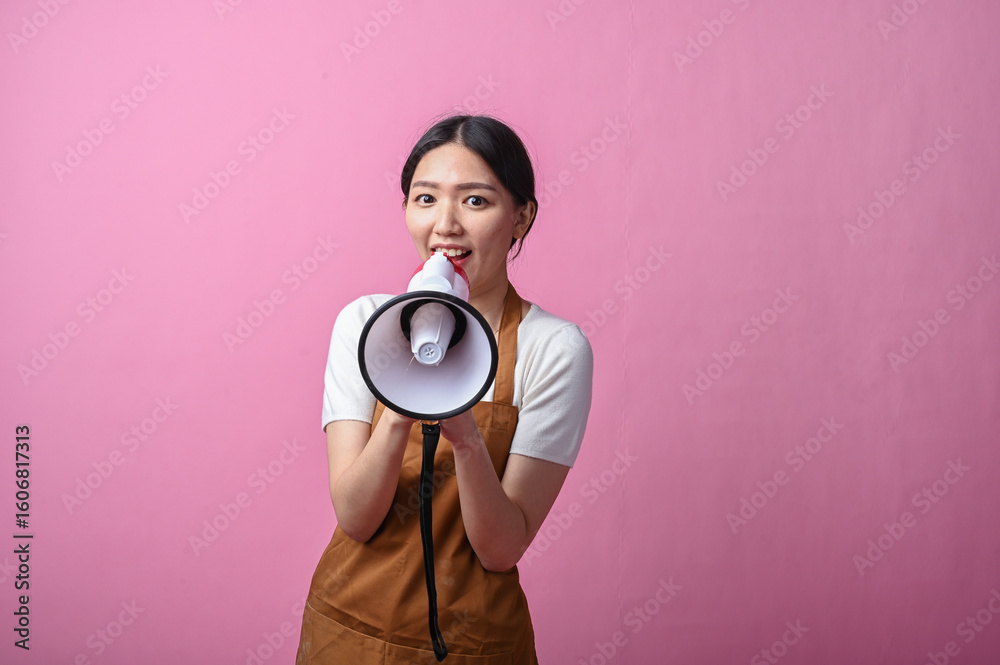Fototapeta premium Asian woman in brown apron holding a white and red megaphone, appearing to speak confidently, standing against a solid pink background