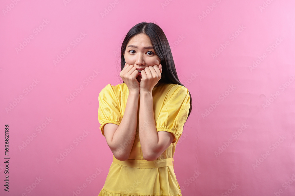 Asian woman in yellow dress looking scared or shocked with clenched fists near her mouth against a pink background. Her wide eyes and posture express surprise or fear in a studio setting Asian woman in yellow dress looking scared or shocked with clenched fists near her mouth against a pink background. Her wide eyes and posture express surprise or fear in a studio setting