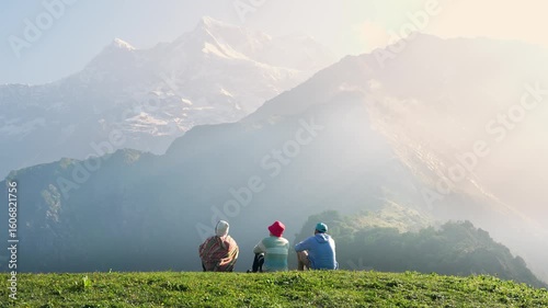 Young men sit on the edge of a mountain, taking in the view of the valley with trees, blue sky, and surrounding mountains along the Madmaheshwar Temple trek, near Ransi Village, Uttarakhand, India.