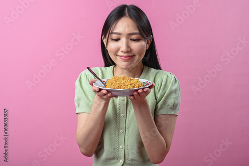 Asian woman in green top laughing joyfully while holding a forkful of instant noodles above a plate, in front of a vibrant pink background