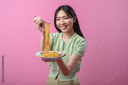 Asian woman in green top smiling while lifting a forkful of cooked instant noodles from a plate, against a plain pink background