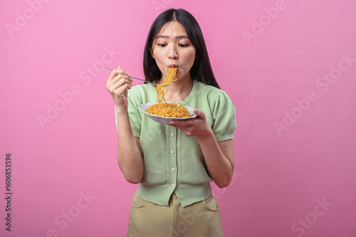 Asian woman in green top eating instant noodles with a fork from a white plate, enjoying her food against a vibrant pink background