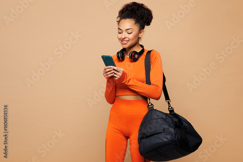Fototapeta Young fitness trainer instructor sporty woman sportsman she wear orange clothes spend time in home gym hold bag use mobile cell phone isolated on plain beige background