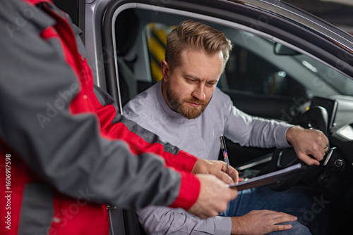 A man seated inside a car reviews and signs documents provided by an automotive professional wearing red overalls in a workshop.