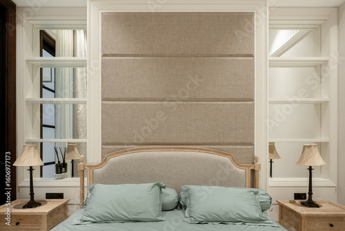 Symmetrical view of a classic bed with an upholstered headboard and a larger fabric wall panel. Flanked by matching wooden nightstands and lamps.