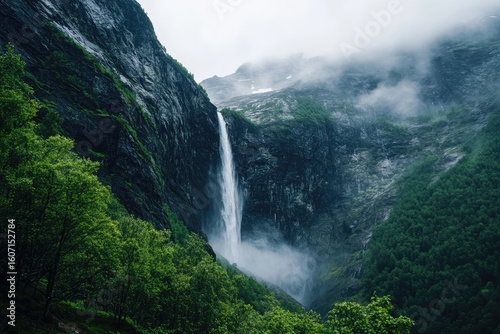 Majestic waterfall cascading down a rocky mountain face, framed by lush green foliage and misty, cloudy mountain peaks