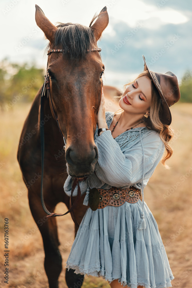 Obraz premium photo real person cowgirl with horse walking on dirt road, field nature summer sky. Woman rider in leather hat holds dark brown stallion by reins sexy Girl blue short dress high boots wild west style
