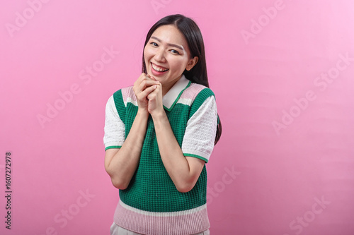 Asian woman smiling sweetly with both hands clasped under her chin, expressing happiness and warmth against a bright pink background