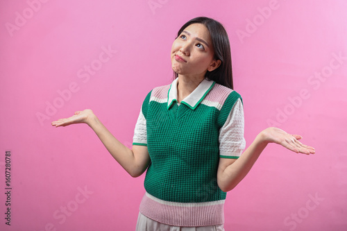 Asian woman shrugging with confused expression, wearing green and pink sweater vest, arms raised, against solid pink background in studio