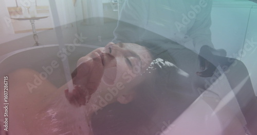 Reclining woman feeling water flowing from faucet over brow in spa room, with bathtub, chrome taps