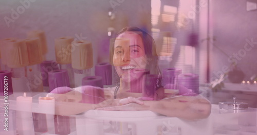 Leaning woman wearing swimsuit top in bathtub in spa room, with candles, towels, window, plant