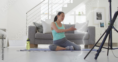 Stretching Asian woman wearing tank top, leggings on mat in living room, with water bottle, tripod