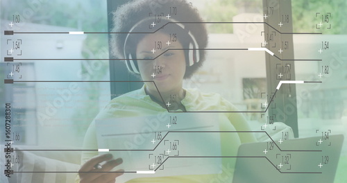 African American woman wearing headphones analyzing data on laptop scrolling phone on couch at home