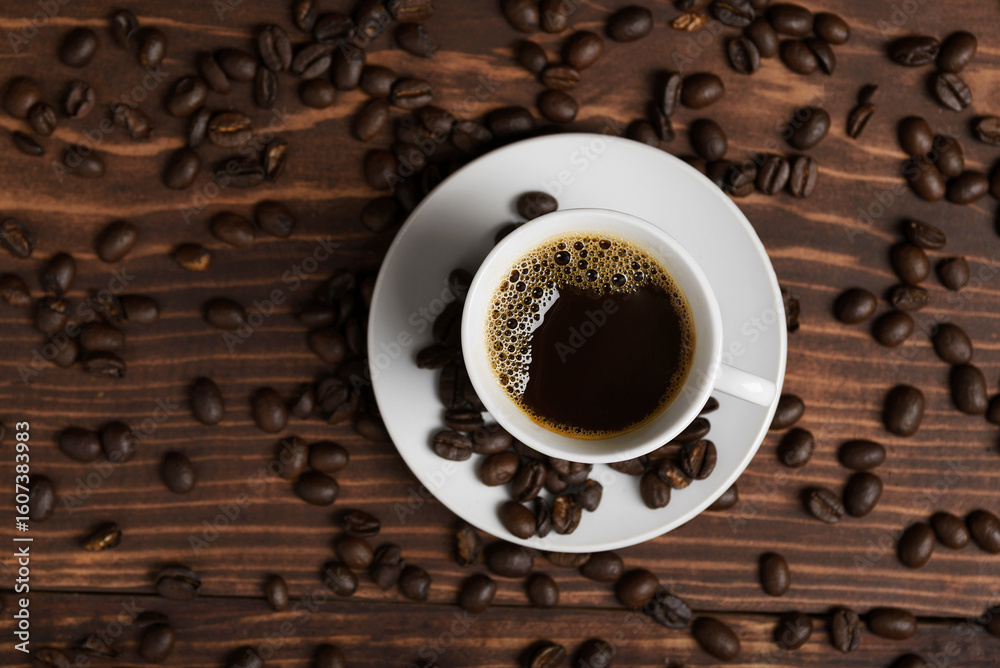 Fototapeta premium A cup of dark roasted coffee on a white saucer, surrounded by coffee beans on a brown wooden surface