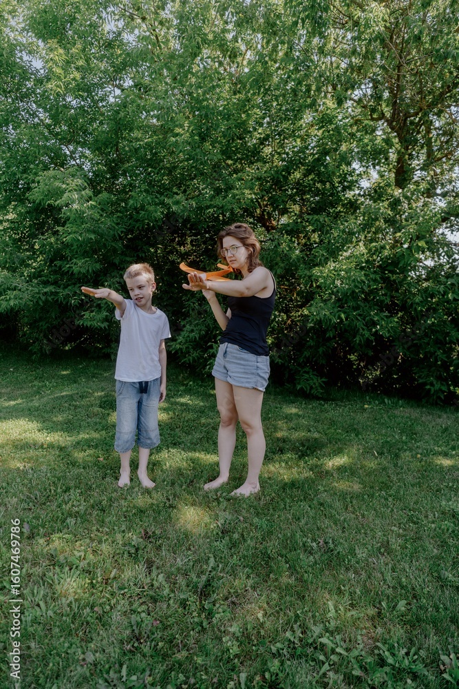 Obraz premium A mother and her son with hearing aids joyfully launch a paper airplane in a sunlit park, capturing a heartwarming moment of inclusive play and bonding in nature