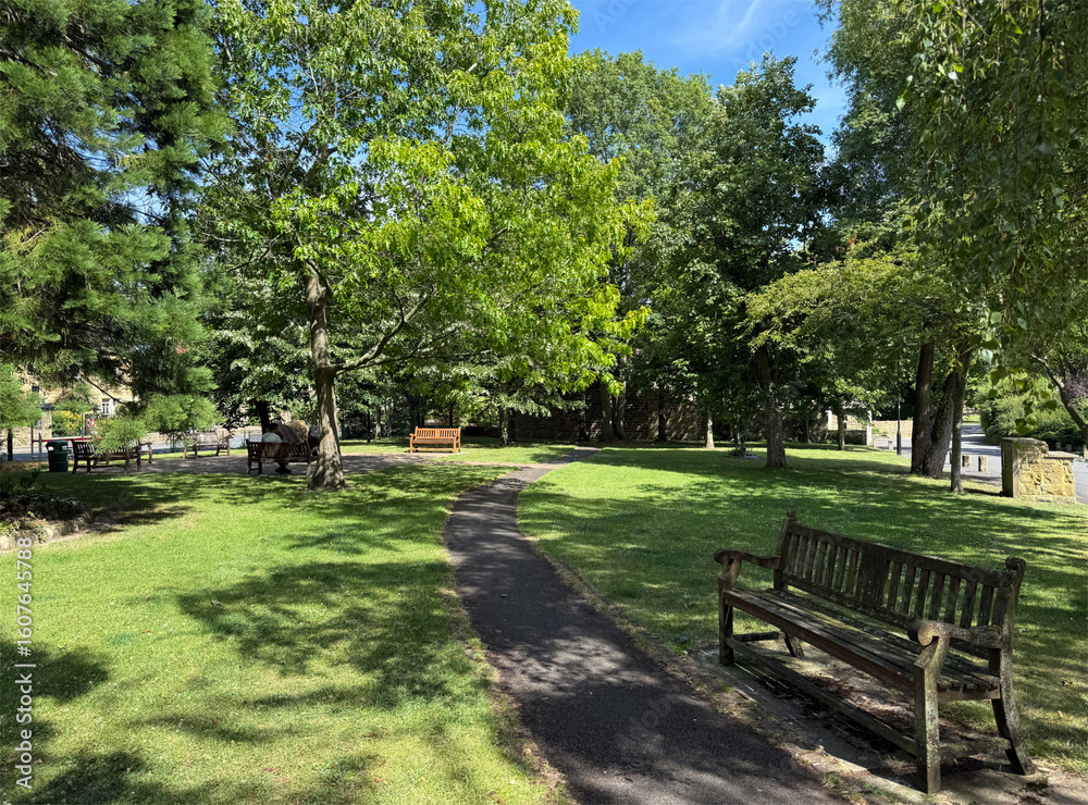 Obraz premium Sunny park path through green shade and benches. A tranquil play of light and leaf in, Burley-in-Wharfedale, Ilkley, UK. 