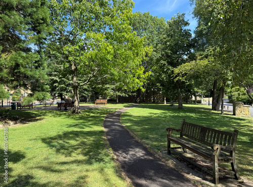 Fototapeta Naklejka Na Ścianę i Meble -  Sunny park path through green shade and benches. A tranquil play of light and leaf in, Burley-in-Wharfedale, Ilkley, UK. 