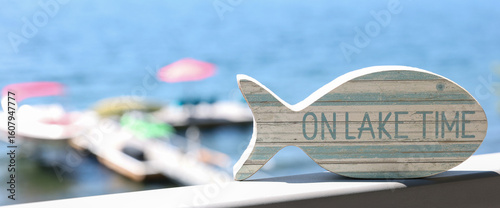 A small fish shaped wooden sign that says On Lake Time outside on a hot summer day near the water. 
