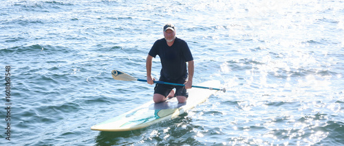 Happy middle aged man resting on his knees on a paddle board outside on a lake. He is holding a paddle over the water and working to maintain balance. 