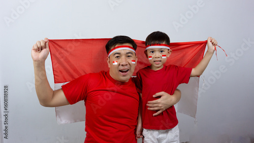 Asian father and son with happy and cheerful expressions wearing red tops and headbands while holding Indonesian flags, isolated on white background. Indonesian Independence Day concept.