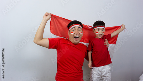 Asian father and son with happy and cheerful expressions wearing red tops and headbands while holding Indonesian flags, isolated on white background. Indonesian Independence Day concept.