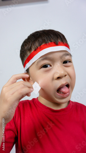 Close-up portrait of an Asian boy wearing a red top and trying remove sticker on his face, isolated on a white background. Indonesian Independence Day concept.