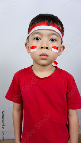 Close-up portrait of an Asian boy wearing a red top and an Indonesian flag ribbon on his forehead and a sticker on his face, isolated on a white background. Indonesian Independence Day concept