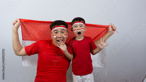 Asian father and son with happy and cheerful expressions wearing red tops and headbands while holding Indonesian flags, isolated on white background. Indonesian Independence Day concept.