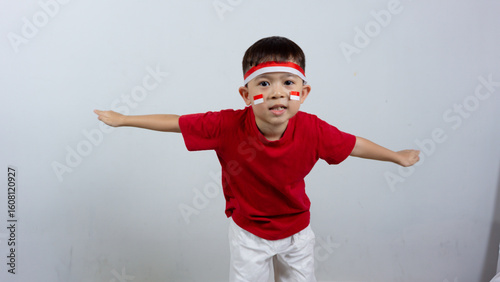 A cheerful Indonesian boy wearing a red top and headband, a mini Indonesian flag sticker on his face, posing like an airplane and smiling. Indonesian Independence Day concept