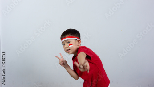Happy Indonesian boy wearing red top and headband, miniature Indonesian flag sticker on his face, posing peacefully with his finger, isolated on white background. Indonesian Independence Day concept