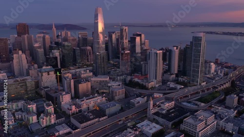 Aerial view of San Francisco, California, USA, at dusk. Skyscrapers dominate the skyline, reflecting the setting sun. Cars travel on the highway below.