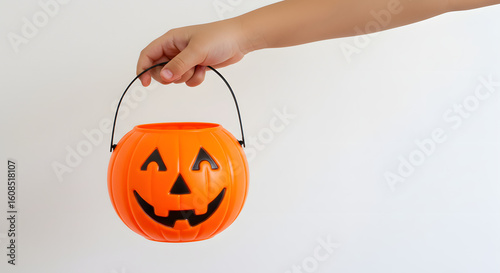 A child's hand holding a festive orange jack-o'-lantern pumpkin bucket for Halloween trick-or-treating against a white background.