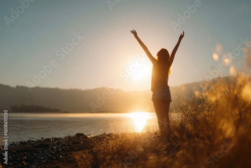 Woman standing with arms raised in celebration by a tranquil lakeside at golden hour, surrounded by dry grass and soft sunlight, embracing nature, freedom, and inner peace