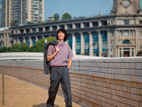 Portrait of handsome Chinese young man with black curly hair wearing stripe shirt posing with suit on shoulder with urban background. Male fashion, cool Asian man lifestyle.