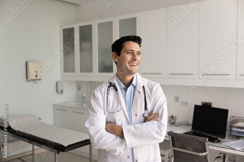 Happy joyful Hispanic doctor man standing in hospital examination room with arms crossed in confident gesture, looking away with toothy smile, laughing. Young physician, general practitioner portrait