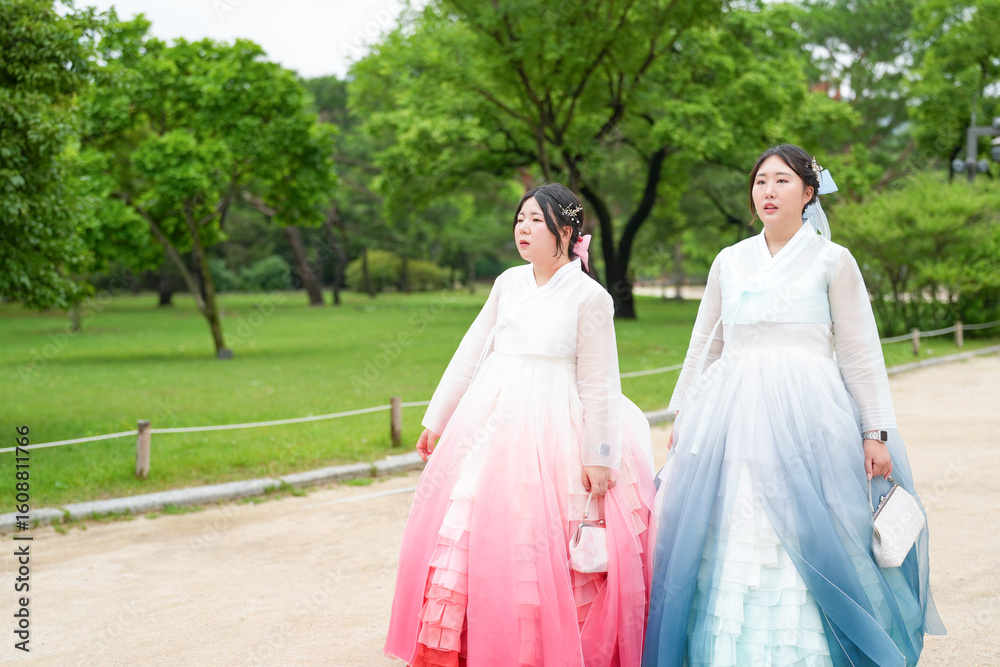 Fototapeta premium Two Korean women in their twenties stroll through a lush green park at Gyeongbokgung Palace, wearing cute pink and sky-blue hanbok, smiling and chatting beneath a clear summer sky. Seoul, June.