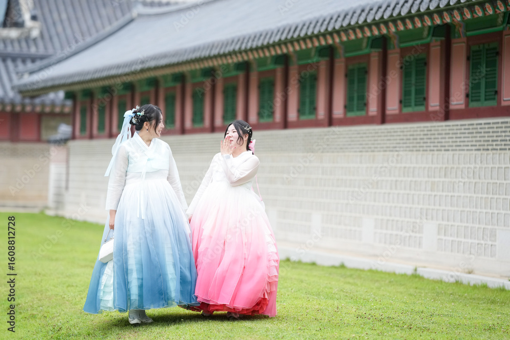 Obraz premium Two Korean women in their twenties walk across a grassy lawn before a historic hall at Gyeongbokgung Palace, wearing cute pink and sky-blue hanbok under a clear summer sky peacefully. Seoul, June.