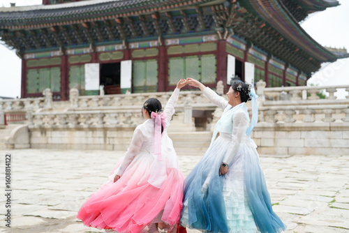 Two Korean women in their twenties dance gracefully in a sunlit plaza at Gyeongbokgung Palace amid historic architecture, wearing cute pink and sky-blue hanbok under a clear summer sky. Seoul, June.