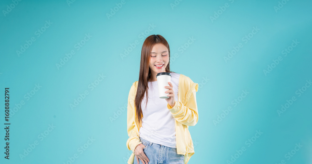 Relaxed young Asian woman enjoying the aroma of coffee while standing against a blue background, representing morning energy, lifestyle, refreshment, and positive vibes. Relaxed young Asian woman enjoying the aroma of coffee while standing against a blue background, representing morning energy, lifestyle, refreshment, and positive vibes.