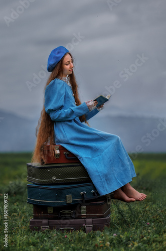 Dreamy vintage girl in blue dress reading a book on stacked suitcases in countryside field under cloudy sky