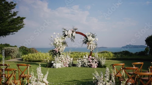 Beautiful outdoor beach wedding setup with floral arch, wooden chairs, and ocean view under blue sky. Romantic, elegant ceremony scene in a tropical garden.