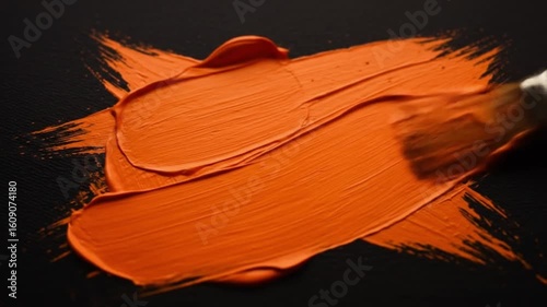 Close up of a paintbrush applying orange paint in a cross shape on a black surface