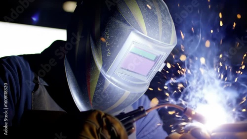closeup on welder at work. intense bright light of torch is contrasted with dark protective helmet, with sparks flying, visually underscoring danger to unprotected eyes. eye injury prevention month