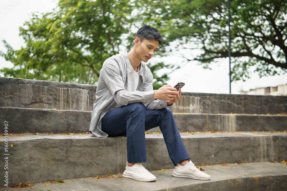 Obraz premium Young asian man sitting on outdoor stone steps, using his smartphone in a peaceful park environment