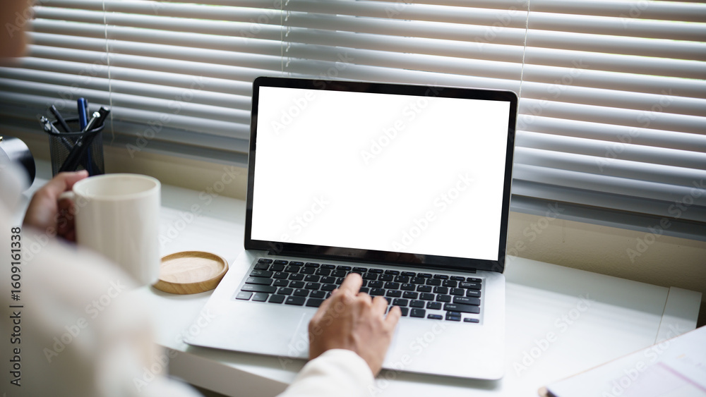Fototapeta premium Close up of woman working on a laptop with blank screen, holding a coffee mug beside a bright window with blinds.