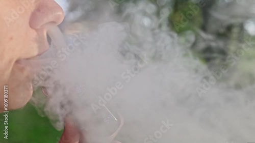 An young woman with red hair vaping in a forested area. The person is captured from the side, exhaling a thick cloud of vapor from an electronic cigarette. Summer landscape in a background.