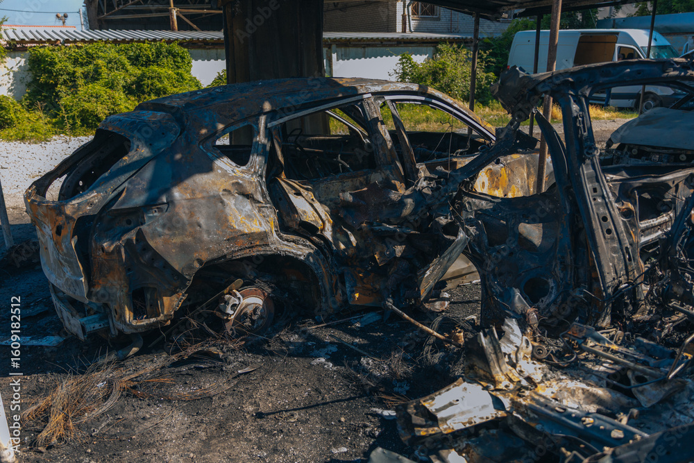Fototapeta premium Burnt carcasses covered in ash and mud show twisted metal and melted structures. Destroyed car interior. Consequences of military conflict and shelling. Destroyed parking lot.