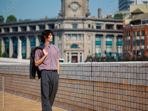 Portrait of handsome Chinese young man with black curly hair wearing stripe shirt posing with suit on shoulder with urban background. Male fashion, cool Asian man lifestyle.