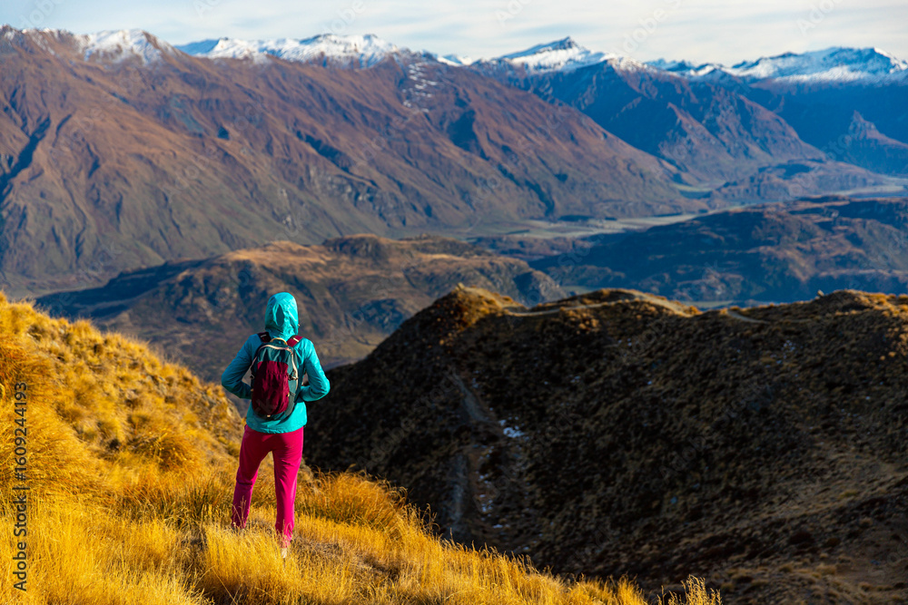 Fototapeta premium Hiker girl hikes along Roys Peak trail and admires stunning panoramic views over Lake Wanaka and surrounding alpine peaks near Wanaka, Otago, South Island, New Zealand.